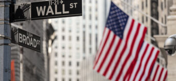 Wall street sign in New York City with American flags and New York Stock Exchange in background.